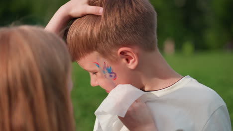 girl gently cleaning paint from brother face with tissue in grassy park, warm light and soft blur background highlighting sibling care and playful bonding during outdoor summer play