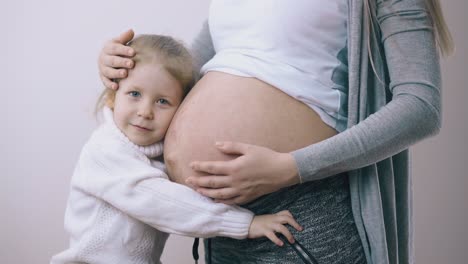 little-kid-hugs-pregnant-mommy-showing-tummy-at-white-wall