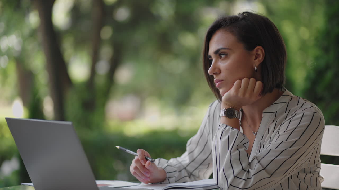 estudiante mujer morena hispana étnica estudiando a distancia a través de internet mientras está sentada en un café de verano con una computadora portátil y escribiendo con un bolígrafo y un cuaderno mientras toma notas en el seminario web