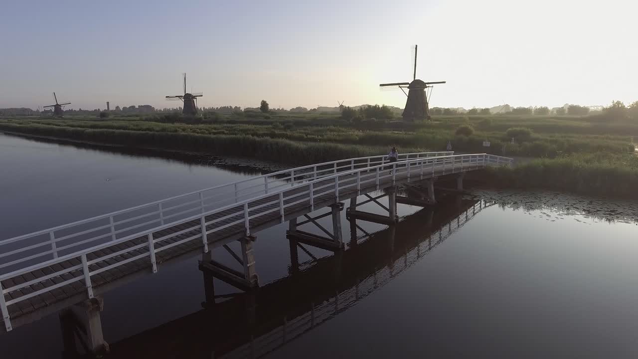 왼쪽으로 패닝하는 드론 샷, 해가 뜨는 동안 네덜란드의 네덜란드 풍차를 바라보며 다리 위를 걷고 돌아선 소녀 주변