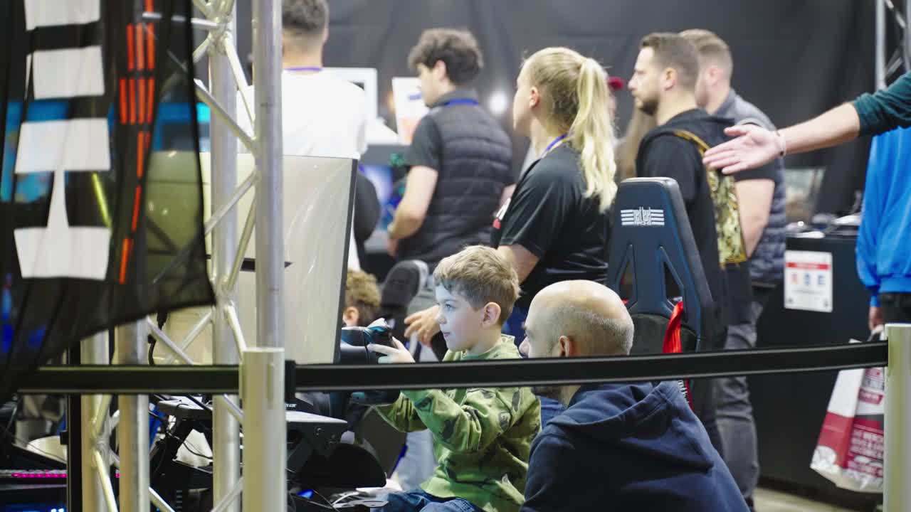Young boy with his father in gaming seat and controller in his hands very concentrated having von while playing new games while female blond instructor is guiding at a convention event LGX Luxembourg