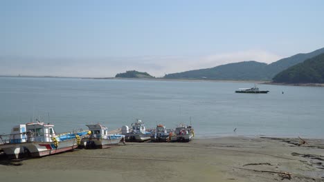 Botes-De-Pescadores-Atrapados-En-El-Barro-Durante-La-Marea-Baja-En-La-Isla-De-Ganghwado-En-Corea-Del-Sur,-Un-Shio-Está-Flotando-En-El-Agua-Del-Mar,-Neblina-Sobre-Los-Picos-De-Las-Montañas,-Boya-De-Navegación-Roja-Flotando-En-El-Mar-Amarillo