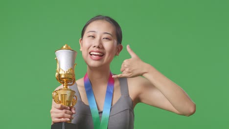 close up of asian woman with a gold medal and trophy showing call me gesture and smiling to camera as the first winner on green screen background in the studio