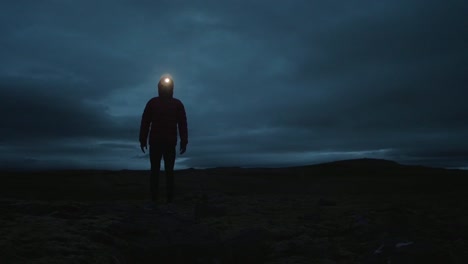 iceland landscape at night, silhouette of a person standing still in frame and looking into the camera with a headlamp on, fast moving clouds in a background