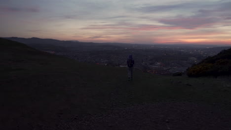 Tiro-Circular-De-Paralaje-De-Una-Niña-Tomando-Una-Foto-Con-Un-Teléfono-Inteligente-Desde-La-Montaña-Arthurs-Seat-En-La-Noche,-El-Atardecer-Con-La-Ciudad-De-Edimburgo-Iluminada-En-El-Fondo-Durante-La-Maravillosa-Hora-Azul