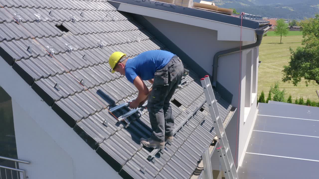Young Caucasian man solar panel roof installer wearing yellow hard hat and blue shirt standing on steep roof of countryside residential home rolling orange wire on spool, static profile