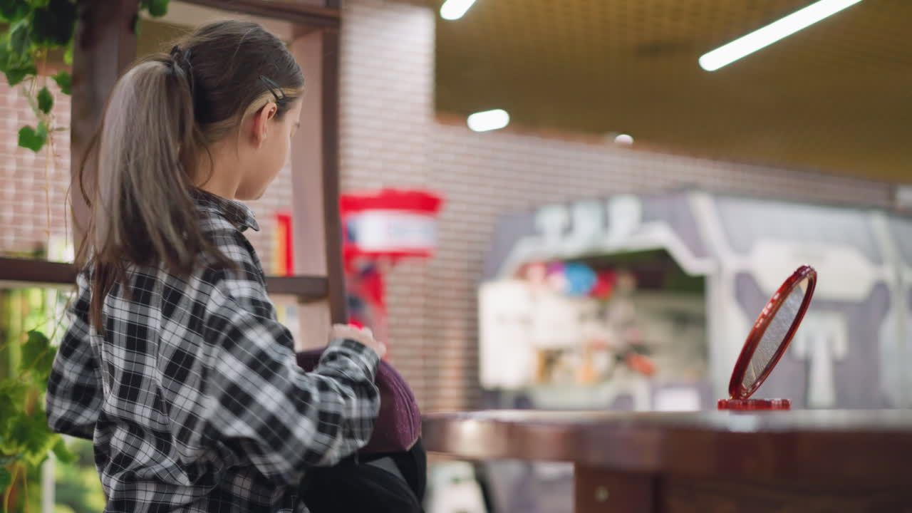 체크색 셔츠를 입은 어린 소녀는 나무 테이블에 앉아 나무 거울을 똑바로 놓고 머리를 부드럽게 조정하면서 그녀의 반사를 바라보고 부드러운 실내 조명과 따뜻한 분위기를 조정합니다.