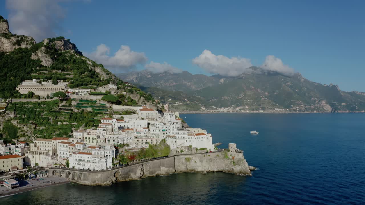 Vista aerea con drone di un'iconica città della Costiera Amalfitana in Italia, con splendidi edifici bianchi a picco sulla scogliera, storico muraglione, spiaggia, profondo Mar Mediterraneo e paesaggio montano.