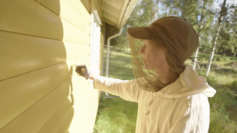 Woman-wearing-mosquito-head-net-paints-wooden-house-with-yellow-paint,-profile