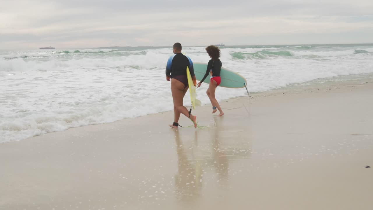Happy african american female friends running into the sea holding surfboards