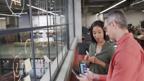 Diverse-male-and-female-colleague-using-tablet-brainstorming-on-glass-wall-in-office,-slow-motion