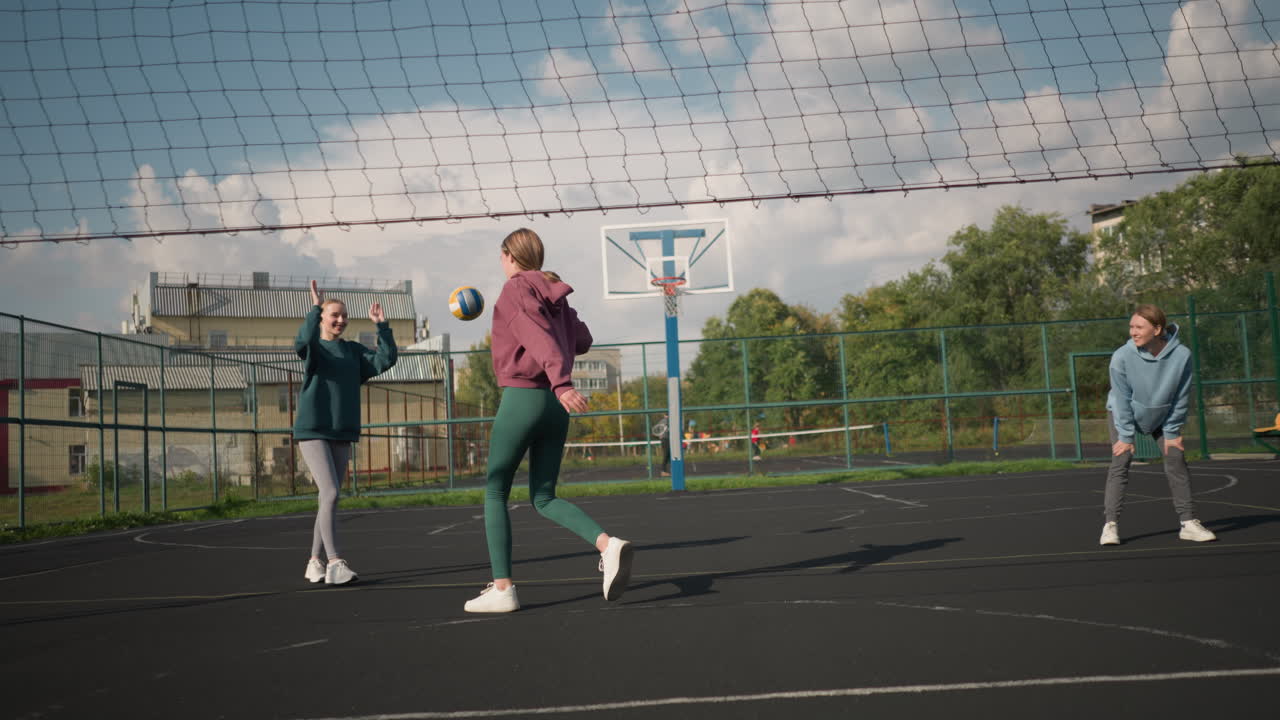 3 명의 배구 선수가 법정에서 공을 놓치고, 두 명의 여성이 놀이를 하면서 포즈를 취하고, 다른 사람들이 바라보는 동안, 녹색과 배경의 건물과 함께 스포츠 경기장에서 활기찬 분위기입니다.