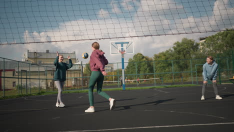 three volleyball players in court miss ball served to them, two ladies pose playfully while other watches, lively atmosphere in sports arena with greenery and building in background