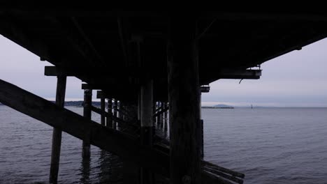 Schwenken-Unter-Pier-Am-Park-Mit-Grauem-Himmel-Und-Wolken-Im-Himmel-über-Dem-Ozeanhorizont-Dunkel-Gruselig-Friedlich