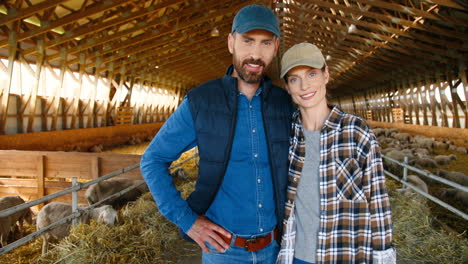 camera zooming on caucasian young couple of farmers looking at camera in a stable with sheep flock