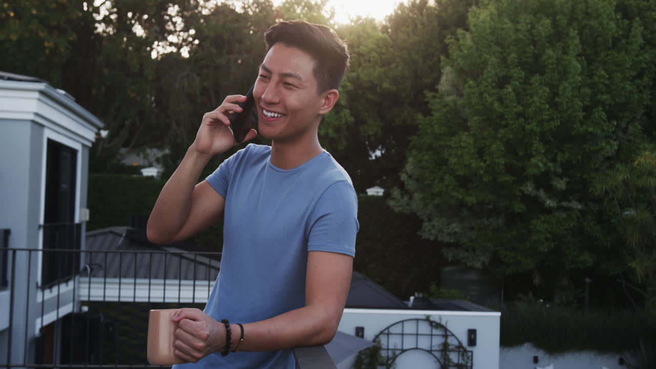 Asian man enjoying phone call outdoors, holding coffee mug, smiling warmly, on balcony, copy space