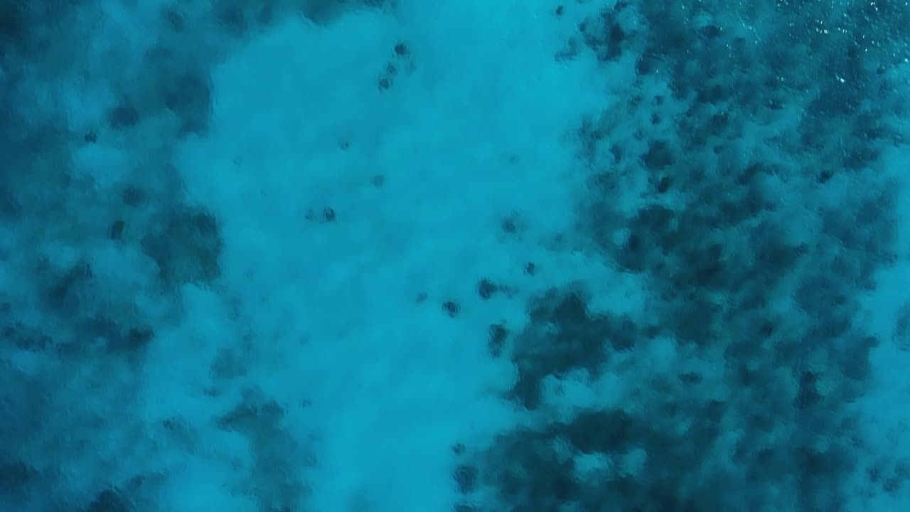 Clear turquoise textured water with sandy bottom and rocky benthos, drone top down
