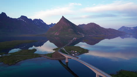 Scenic-drone-shot-of-a-mountain-reflecting-on-the-Surface-with-scenic-brigde-in-Lofoten-during-midnight-sun