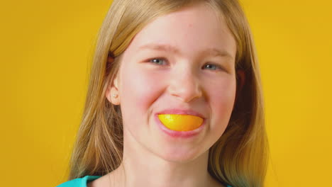 studio portrait of girl using orange segment for mouth and teeth against yellow background