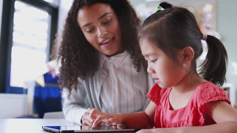 Female-infant-school-teacher-working-with-a-young-Asian-schoolgirl-using-tablet,-close-up,-low-angle