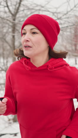 tired adult woman wearing red hoodie and beanie jogging along paved snow covered path outdoors during winter surrounded by barren trees and serene park scenery showing resilience in cold conditions