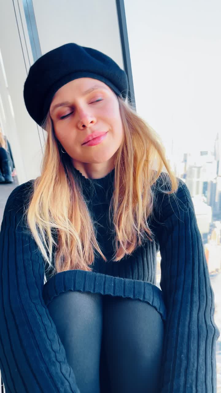 Donna con berretto nero vicino a una finestra di New York. Donna con berretto nero e vestito a coste accanto a una finestra alta con lo skyline sfocato di Manhattan sullo sfondo.
