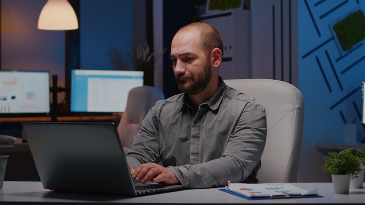 перегруженный работой бизнесмен печатает финансовую статистику на ноутбуке, сидя за столом