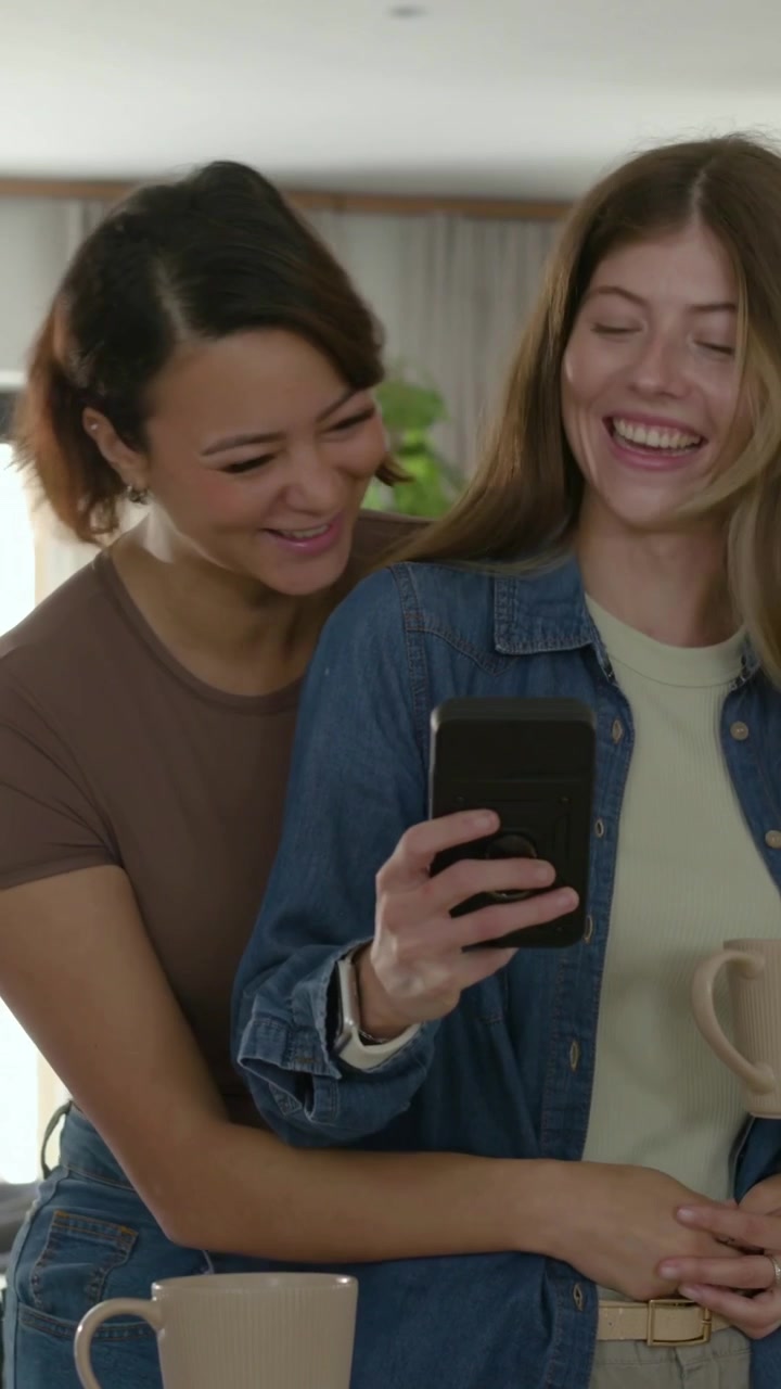 Vídeo vertical: Casal se abraçando, mulher da frente tocando no telefone na cozinha, enquadrando uma selfie, de camiseta marrom jeans.
