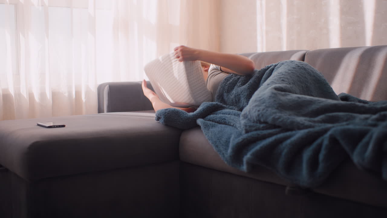 Unhappy girl lying on couch hugging pillow tightly with squinted eyes resisting daylight, wrapped in soft blue blanket as sheer curtains glow in background, avoiding morning start
