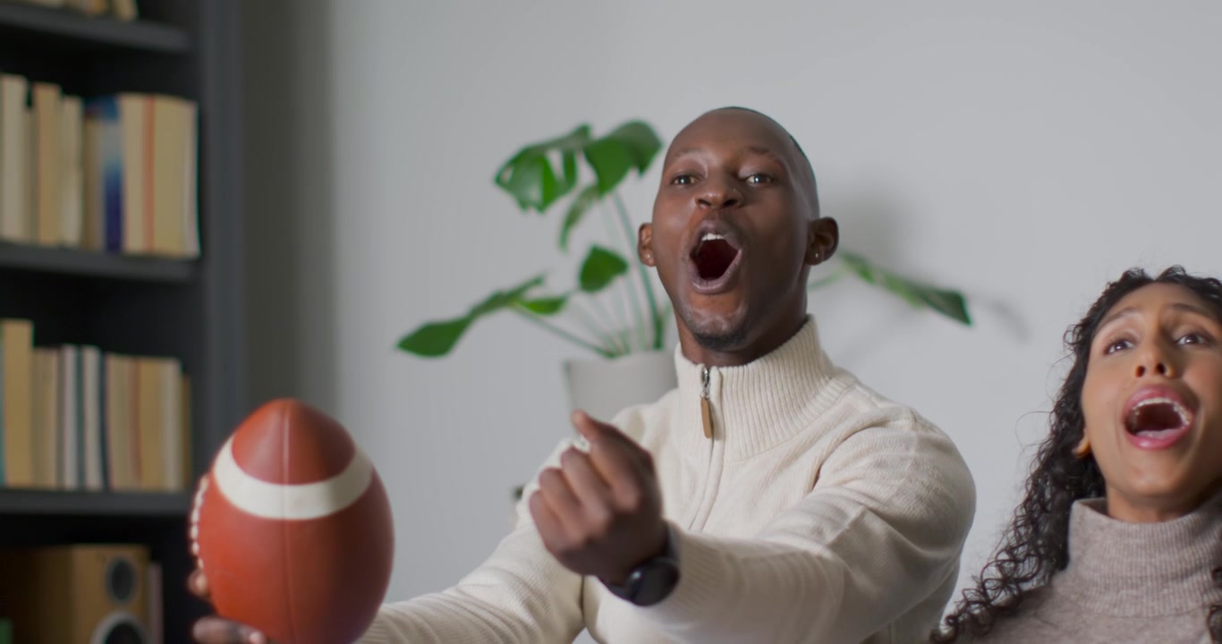 African American couple in living room watching game, man holding football, tossing and reacting
