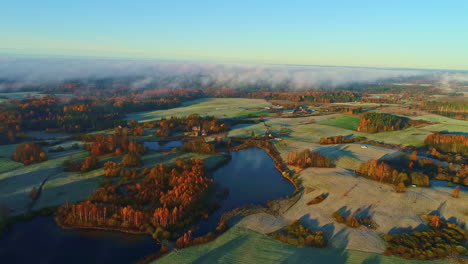 Aerial-drone-shot-flying-over-green-farmlands-beside-small-lakes-on-a-cloudy-day-with-sun-rays-falling-on-the-field