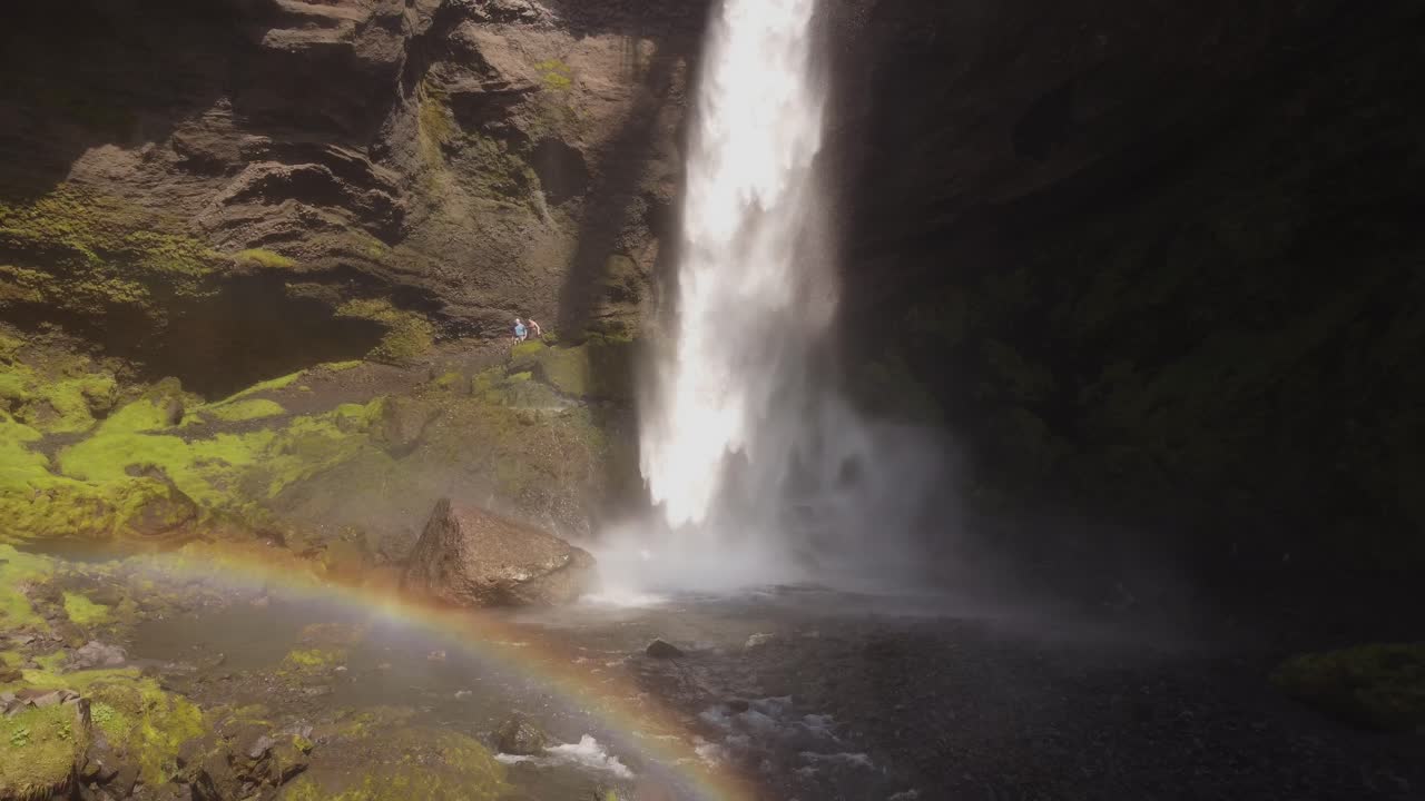 retiro aéreo de un dron de una hermosa cascada en islandia en un día soleado con pájaros volando y un pequeño arco iris frente a los acantilados y rocas verdes de musgo