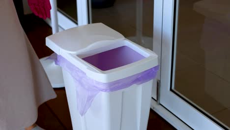 a person wearing gloves disposes of trash in a white bin with a purple liner indoors