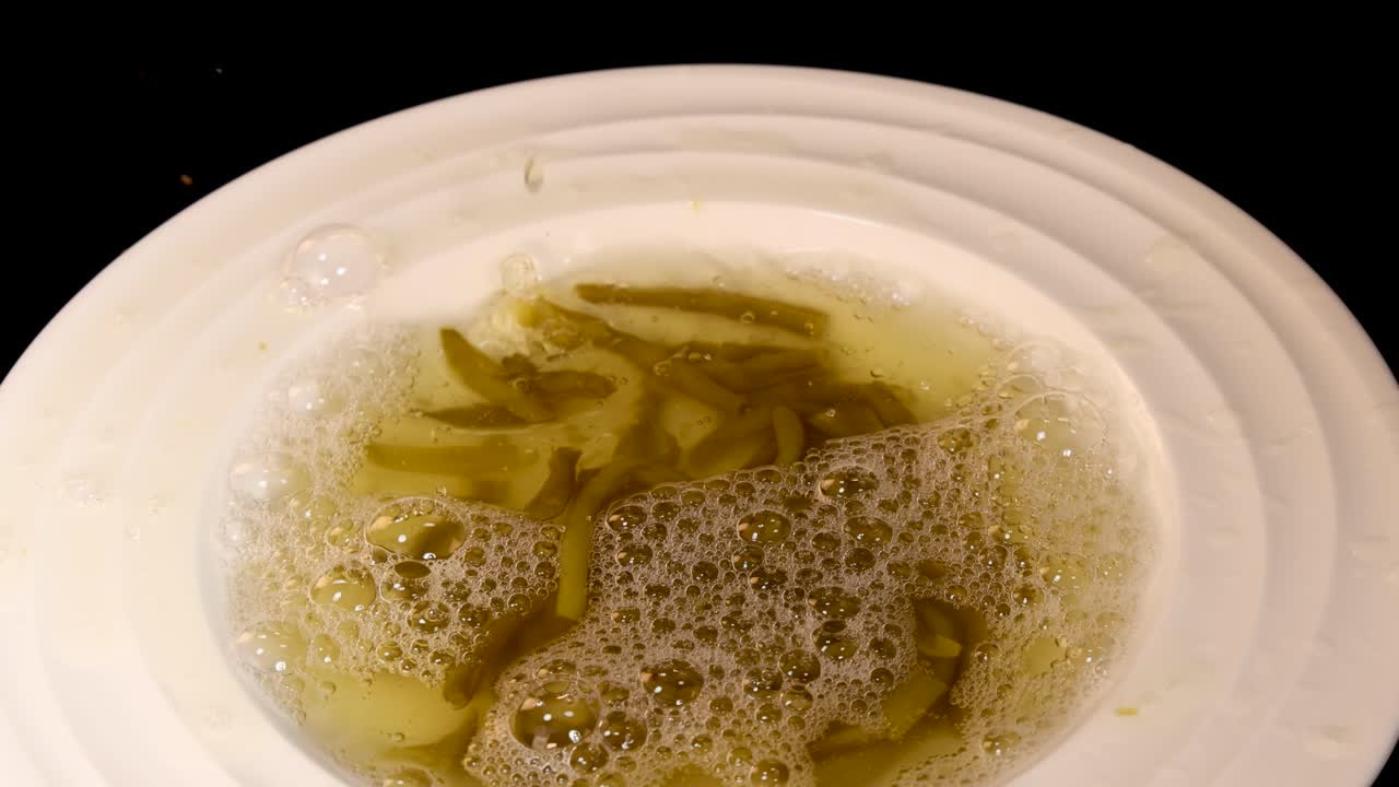Haricot beans and liquid are poured into a white bowl, creating bubbles and movement. Overhead view, even lighting, black background, no visible people