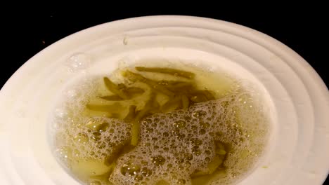 Haricot beans and liquid are poured into a white bowl, creating bubbles and movement. Overhead view, even lighting, black background, no visible people