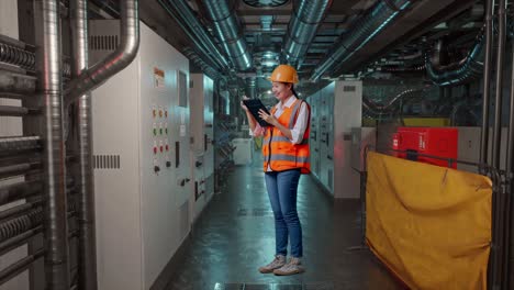 full body side view of asian female engineer with safety helmet taking note on the tablet and looking around while standing in engine control room, work of electrical generators