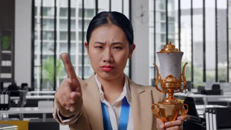 close up of asian business woman in a suit with a gold medal and trophy disapproving with no index finger sign and smiling to camera in the office