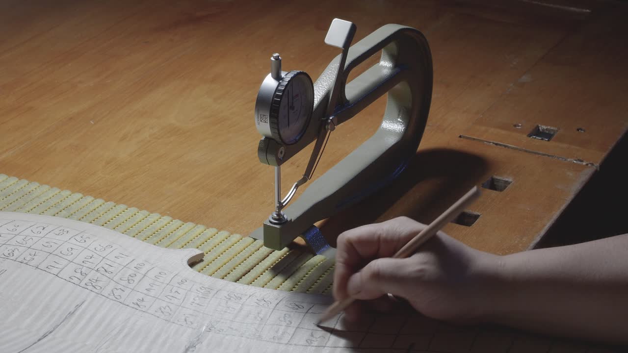Maestro liutaio, costruttore di strumenti a corda, procede con la lavorazione di graduazione sulla tavola posteriore di un violoncello, utilizzando un calibro di spessore per misurare i livelli e una matita per annotare i valori sul legno, nel suo laboratorio a Cremona, Italia.