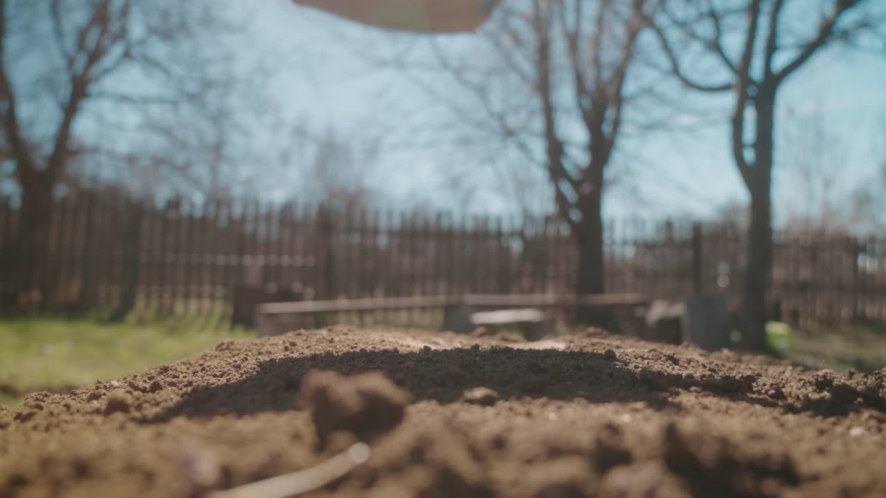Close-up of a shovel preparing soil in a garden on a sunny day with a fence and trees in the background