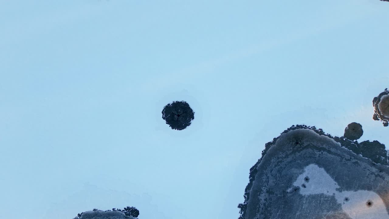 Frozen lake covered in snow with black spots of exposed ice in aerial top-down view