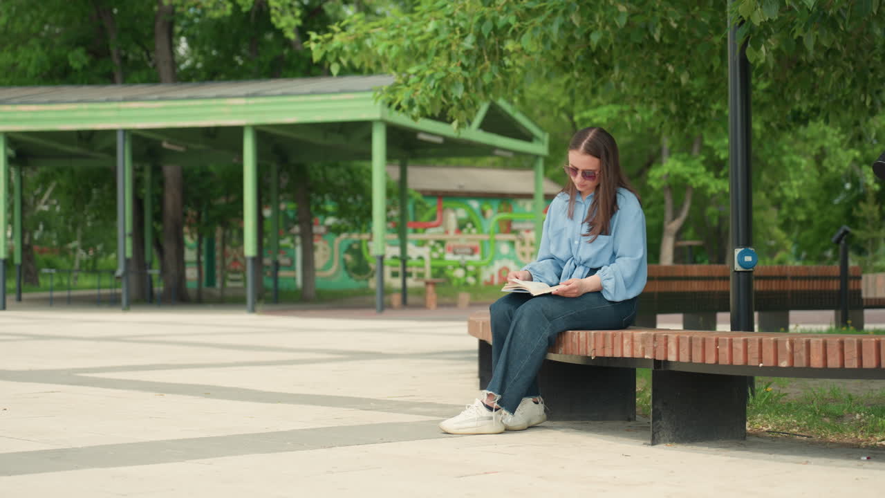 공원 벤치에서 책을 읽고 있는 백인 여성, 데님 재킷과 선글라스를 착용하고 교재에 집중하고 있으며, 펼쳐진 페이지와 노트북이 옆에 놓여 있고, 배경에는 평온한 잎이 무성한 정자가 있으며, 따뜻한 오후 햇살, 고독한 학습 분위기