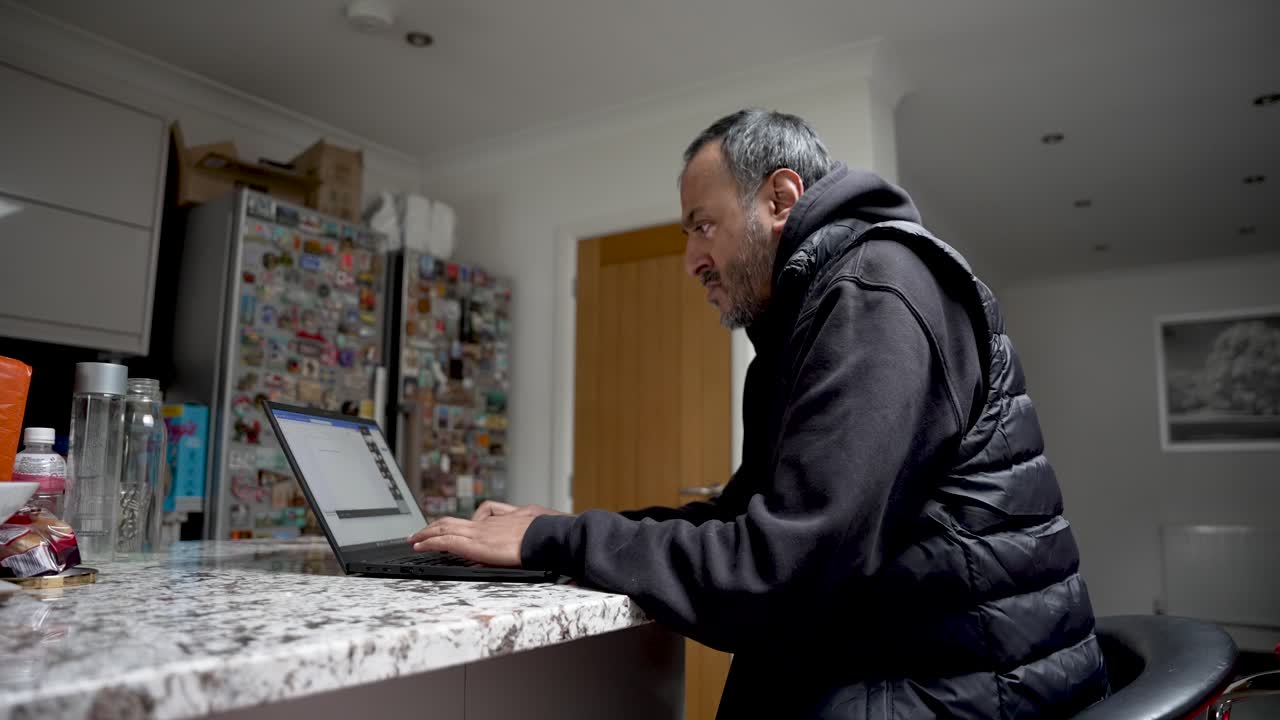 South Asian man working on a laptop at a kitchen table in a home office setting. He is engaged in remote work showcasing a digital nomad lifestyle and flexible working arrangements