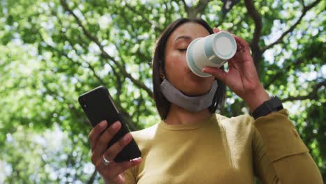 Afroamerikanische-Frau-Mit-Gesichtsmaske-Und-Smartphone,-Die-Im-Park-Kaffee-Trinkt