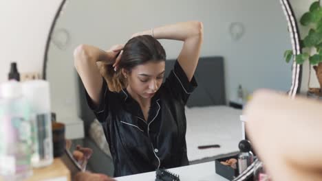 portrait of a young beautiful woman in pajamas sitting in front of the mirror, styling her luxurious hair into a bun, pinning it up, and posing gracefully