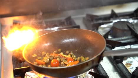 a dramatic scene of flambé cooking, with flames rising above a pan containing cherry tomatoes and capers. the vibrant ingredients are being tossed in a hot pan, creating a mediterranean preparation.