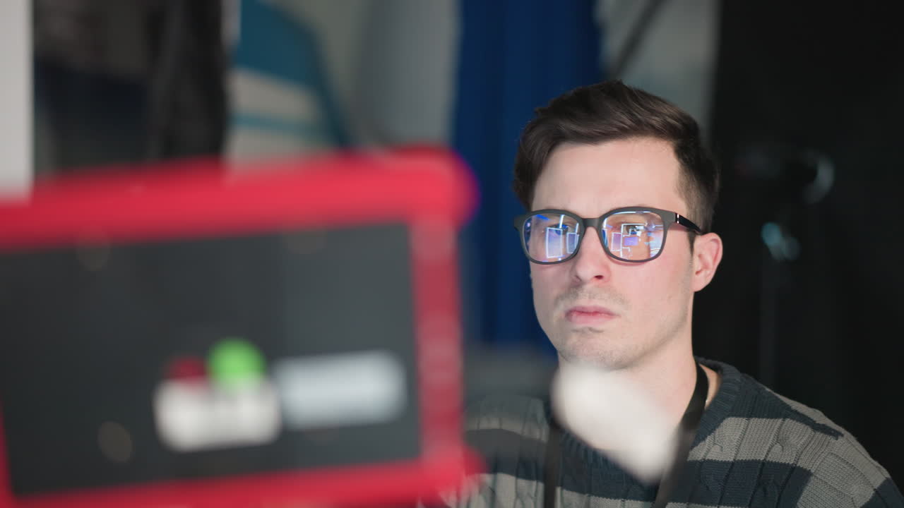 Camera operator with glasses focusing on camera monitor while adjusting equipment in TV studio. Reflections in glasses show camera setup, emphasizing detailed attention during filming process