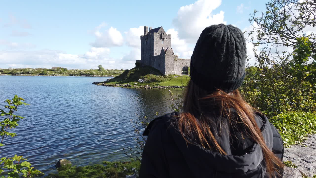 Visitor standing near historic dunguaire castle, overlooking scenic landscape with water reflection under bright summer sky in county galway, ireland