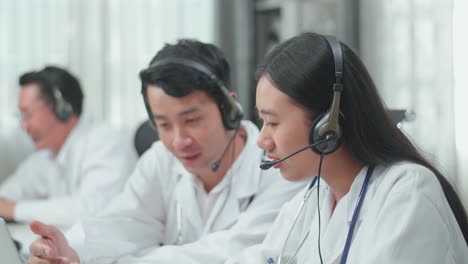 Close Up Of An Asian Man And A Woman Doctors Wearing Headsets Working As Call Center Agent Discussing Work While Their Colleague Is Speaking With Customer On The Call At The Office