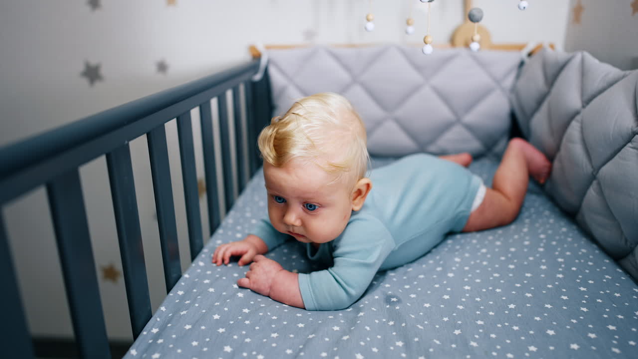 Adorable blond baby boy in blue bodysuit lying in the crib. Calm blue-eyed child looking intently ahead.