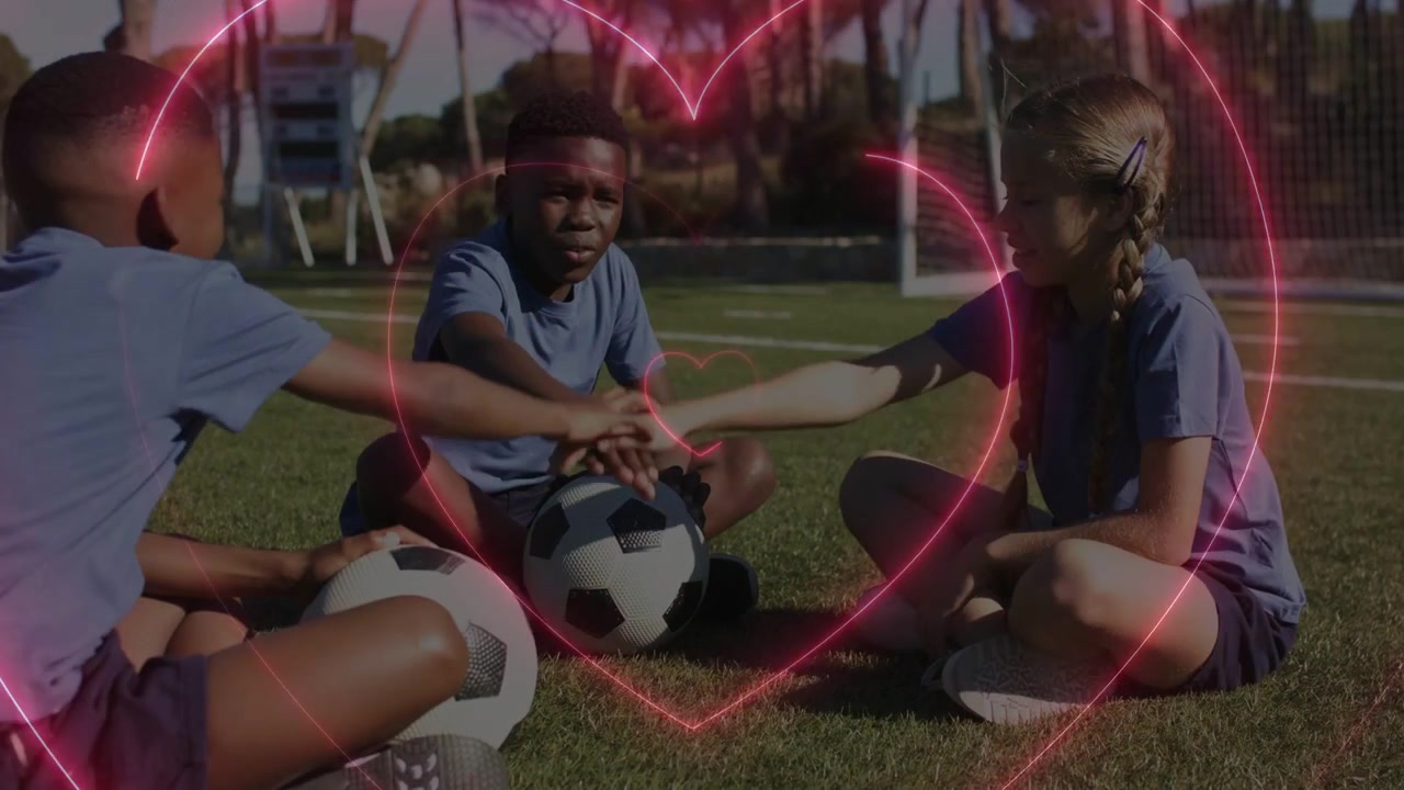 Three kids sitting on pitch, girl reaching stacking hands over balls for sports cheer heart pulsing